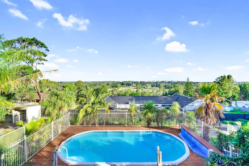 Oval swimming pool with scenic hillside views surrounded by tropical landscaping and blue sky.