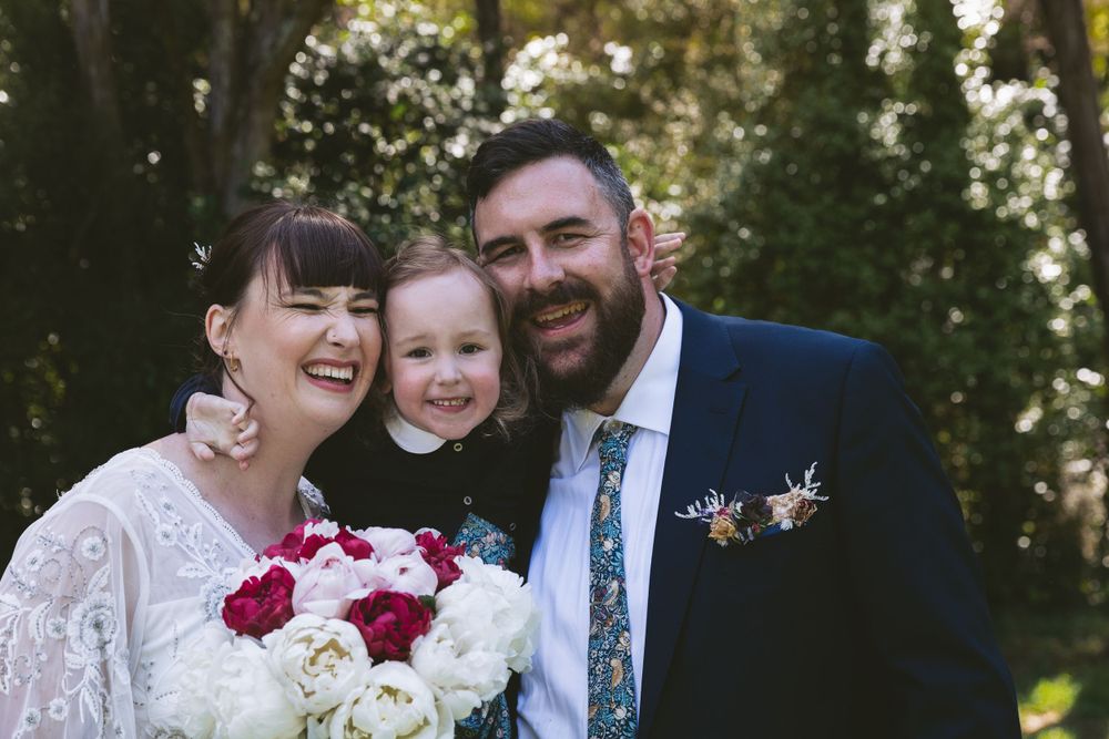 Happy wedding portrait with red and white bouquet against forest backdrop.