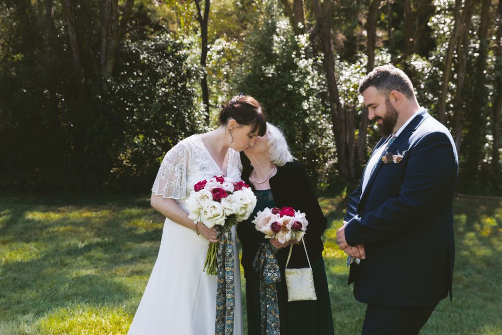 An outdoor wedding ceremony in a wooded setting with a bride in white holding red and white flowers.