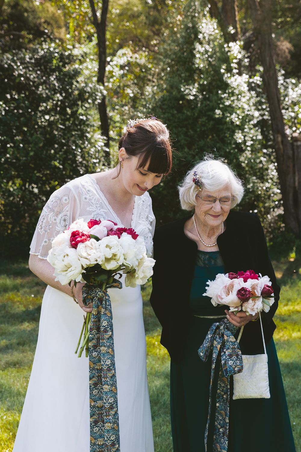 A bride in a white lace dress stands with an elderly woman in black holding matching floral bouquets.
