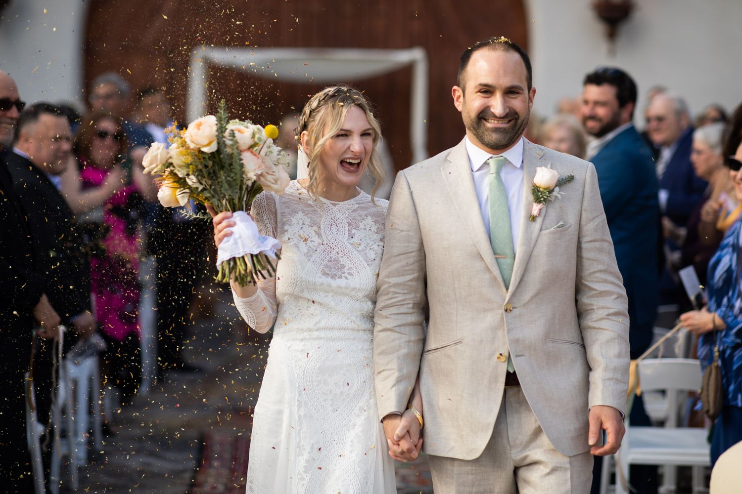 Newlyweds celebrate while walking down the aisle as guests throw confetti in celebration.
