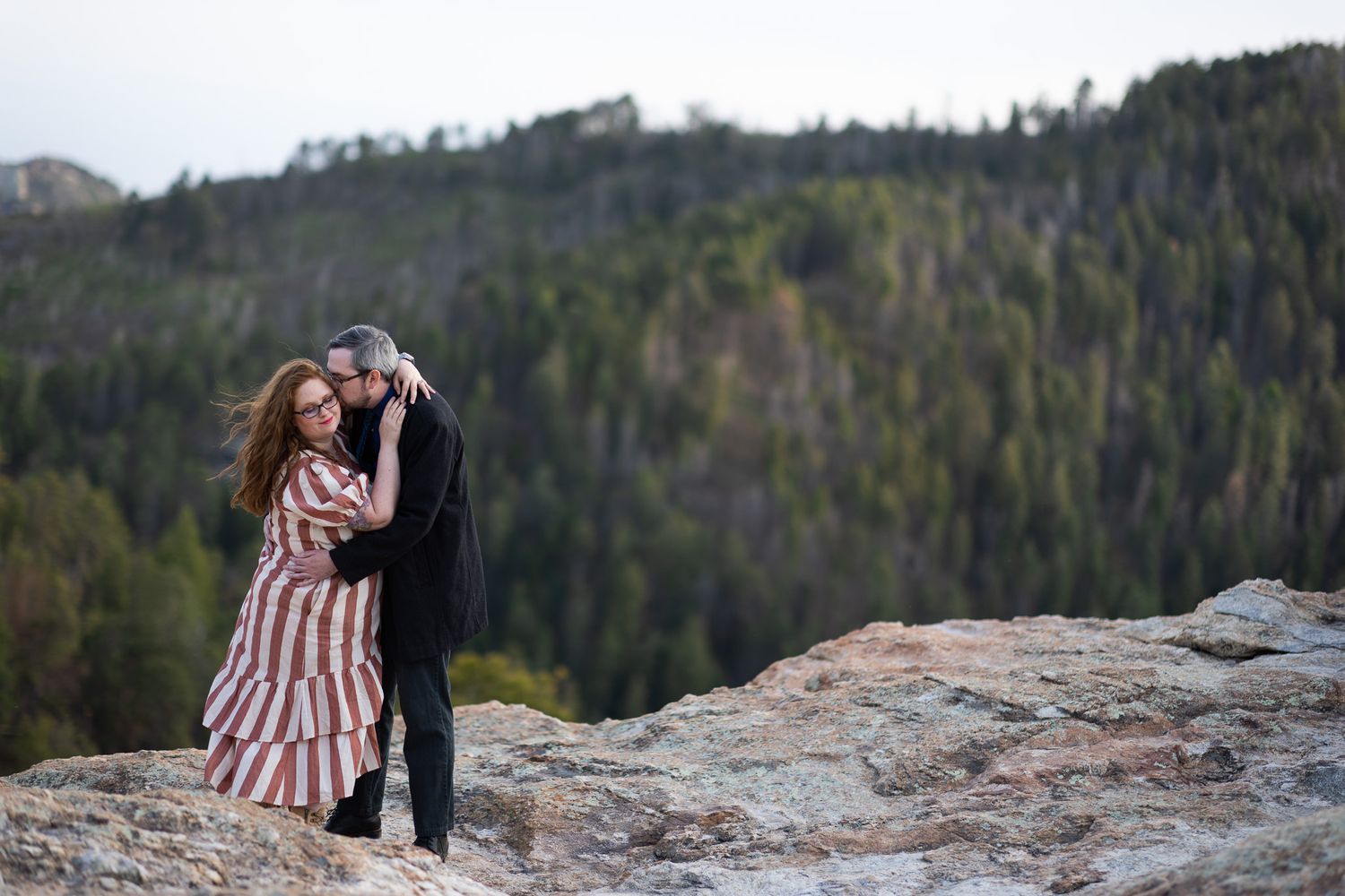 A couple shares an intimate moment on rocky mountain overlook with forest valley below.