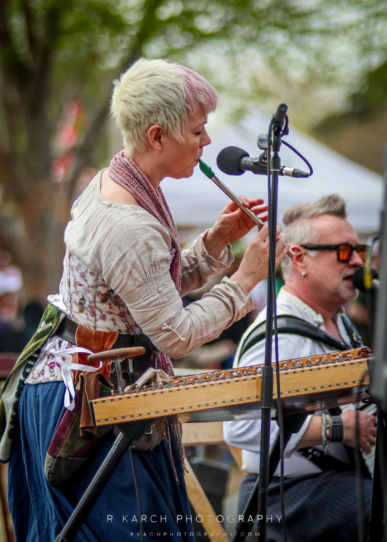 Musician playing dulcimer at outdoor festival with microphone setup.