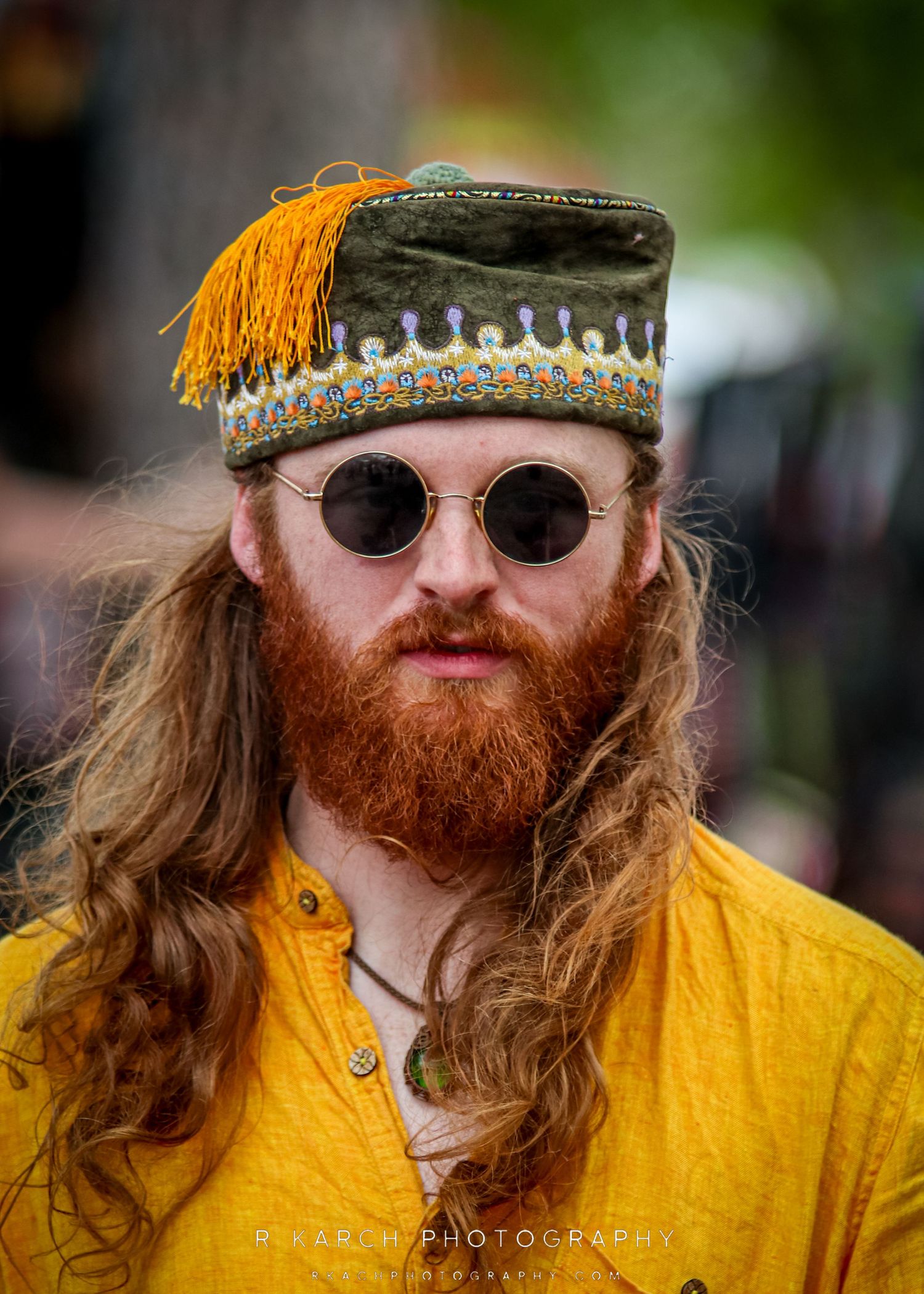 Festival attendee wearing decorative hat with tassel and round sunglasses in bright yellow shirt.