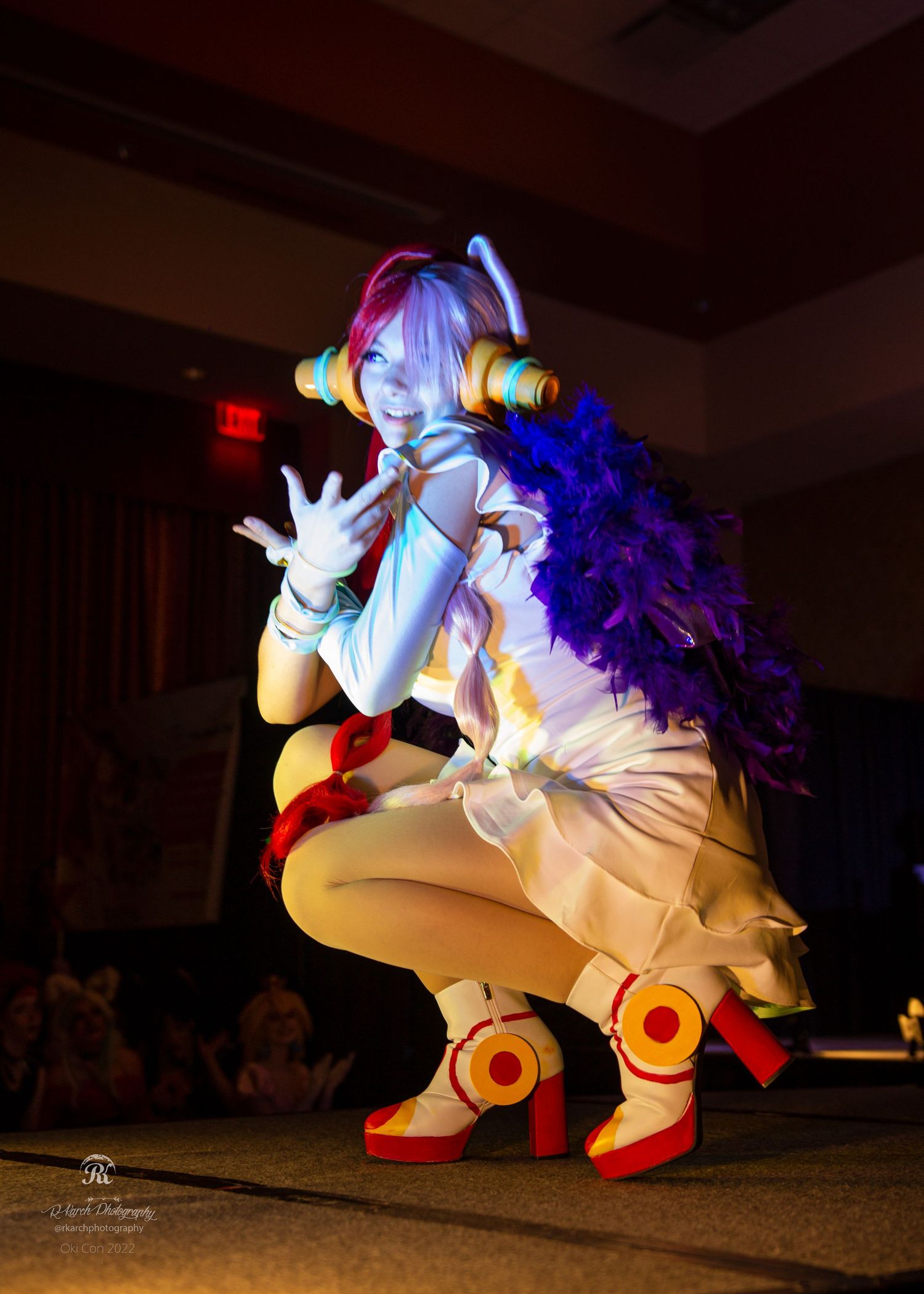 A cosplayer in a vibrant anime-inspired outfit performs on stage with dramatic lighting.