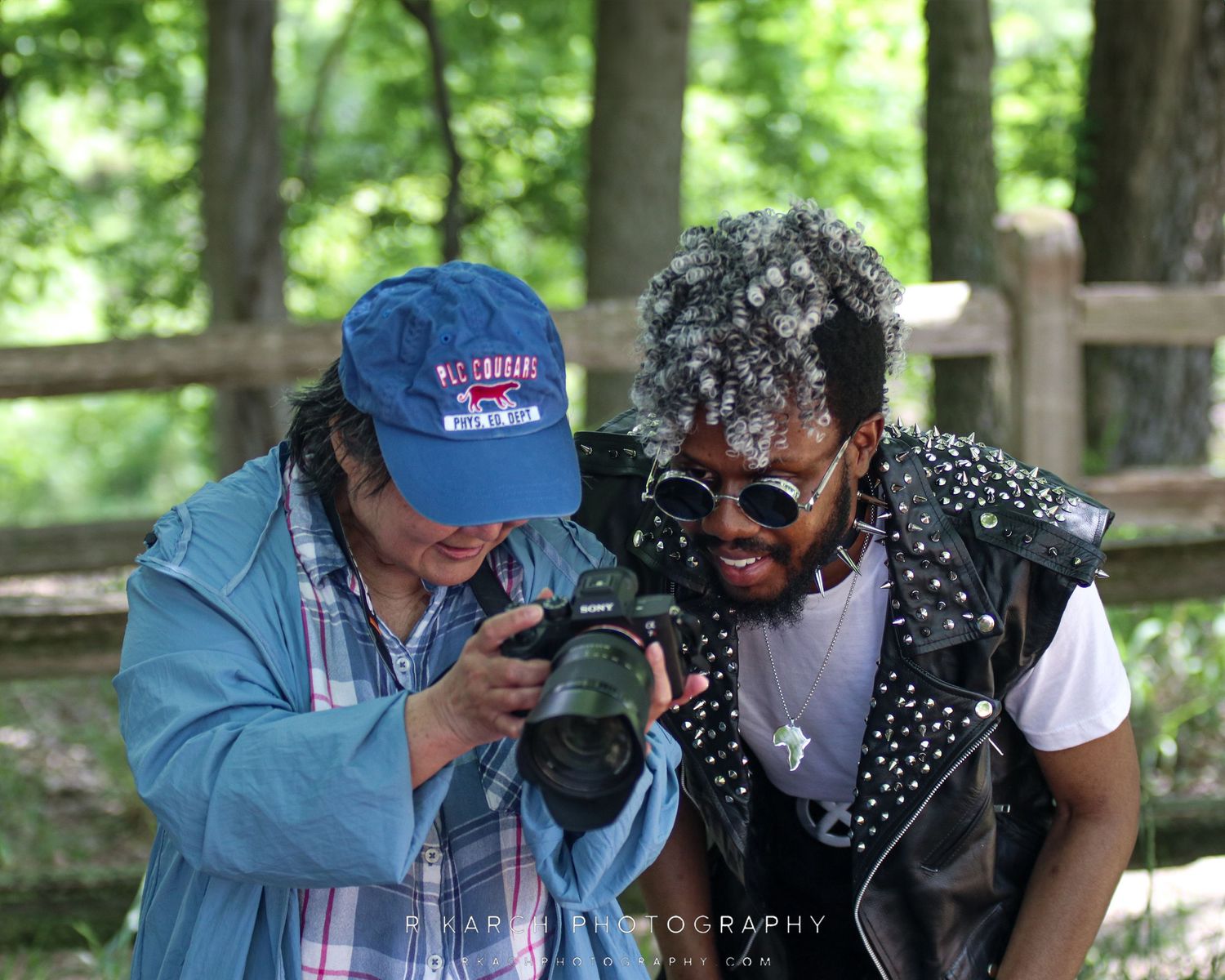 Two event attendees examine a camera together in an outdoor wooded setting.