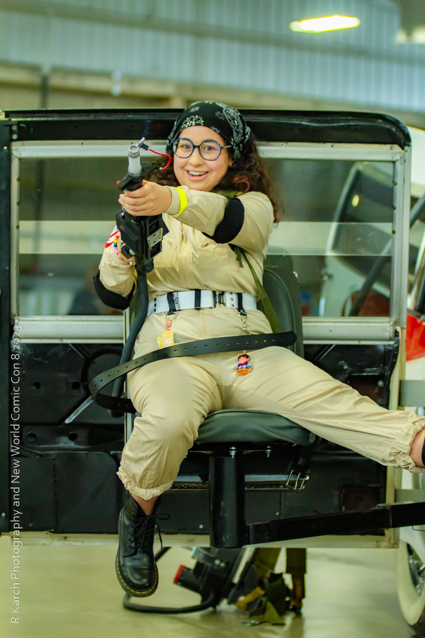 Cosplayer in Ghostbusters uniform poses with prop proton pack in vintage vehicle.