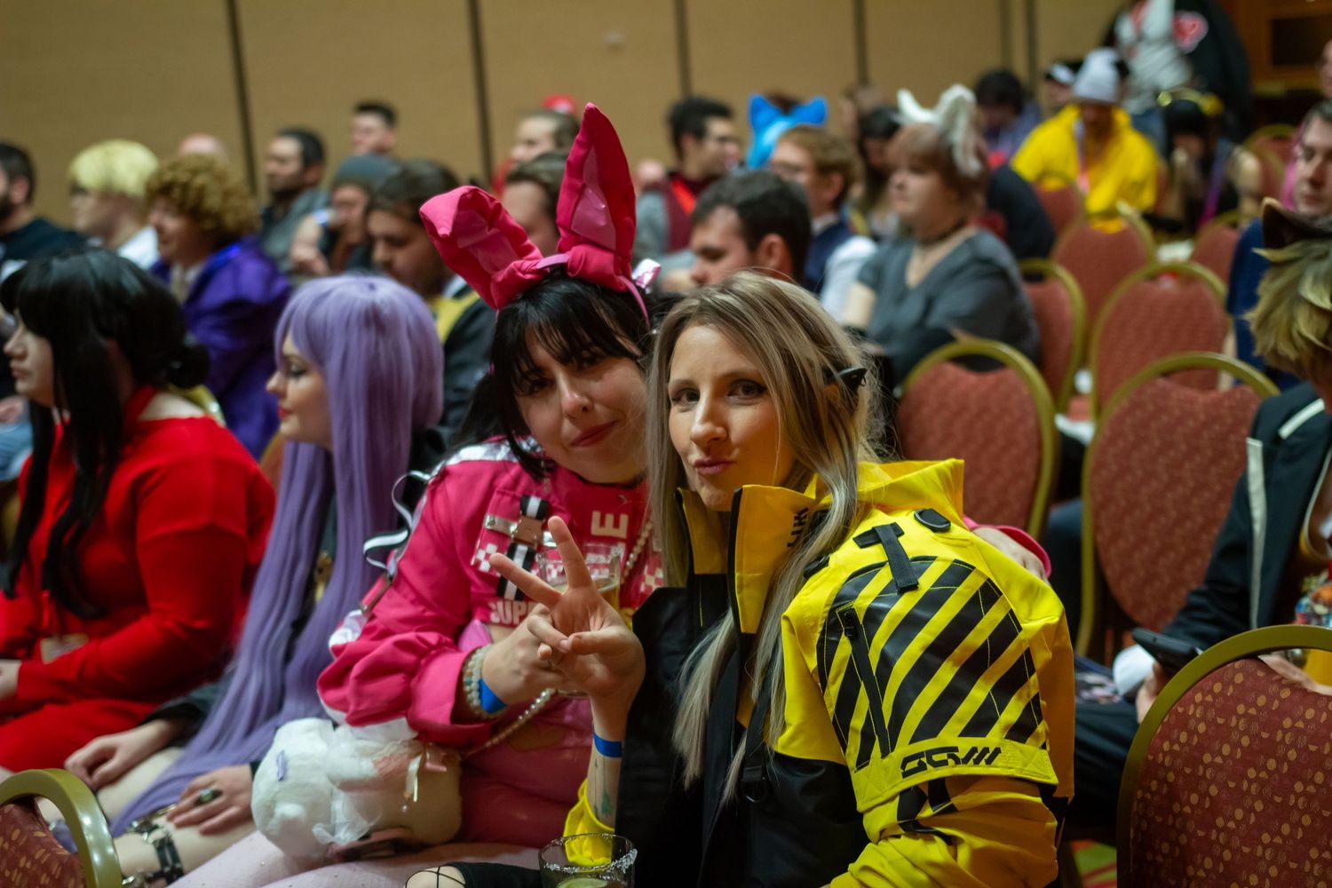 Convention attendees in colorful costumes and wigs seated in a conference hall.