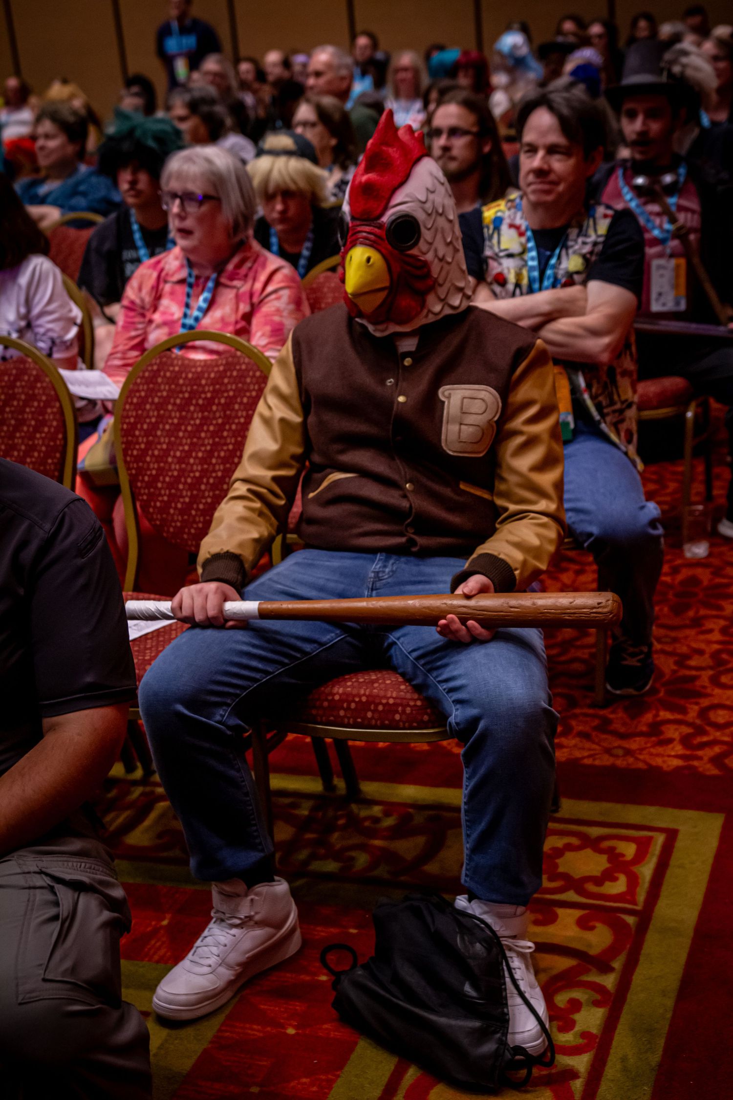 An attendee in a rooster mask sits among the audience at a convention event.