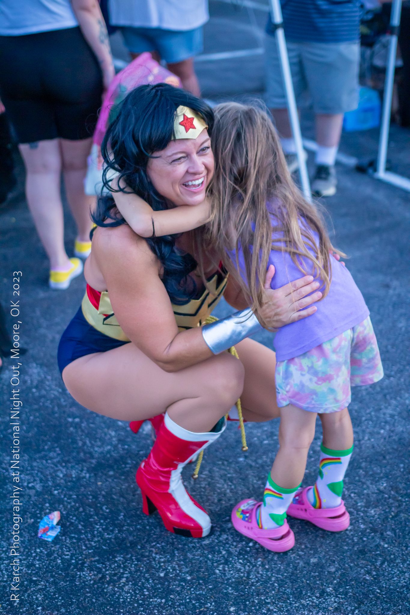 A Wonder Woman cosplayer shares a heartwarming moment with a young fan at an outdoor event.