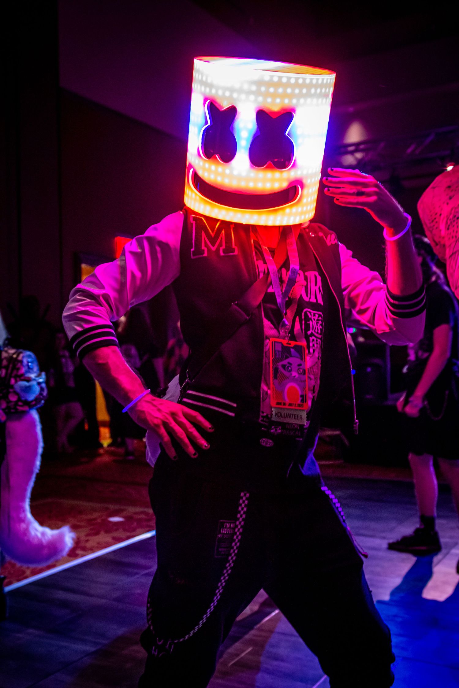 A performer wearing an illuminated Marshmello-style helmet dances on stage in purple lighting.