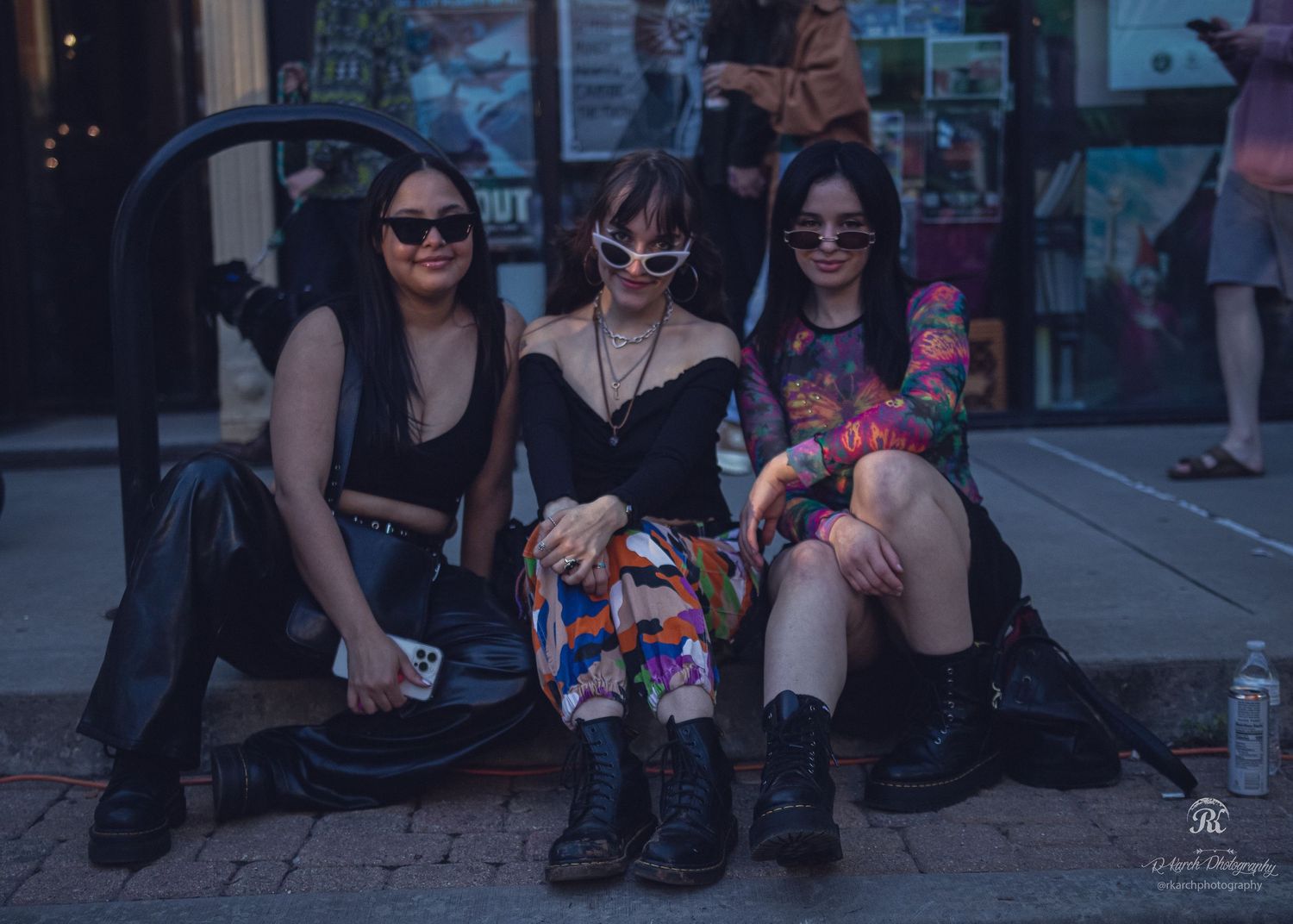 Friends in dark outfits sitting together on a street curb during twilight hours.