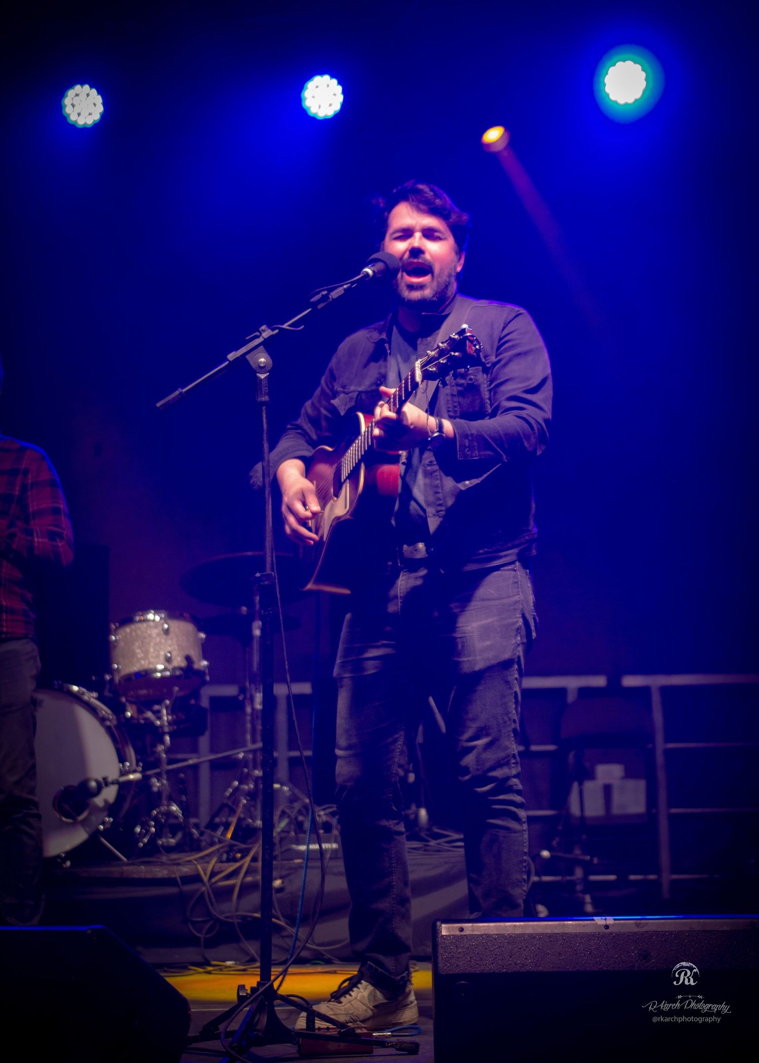 Musician performing with acoustic guitar on stage with blue spotlights and drum kit in background.