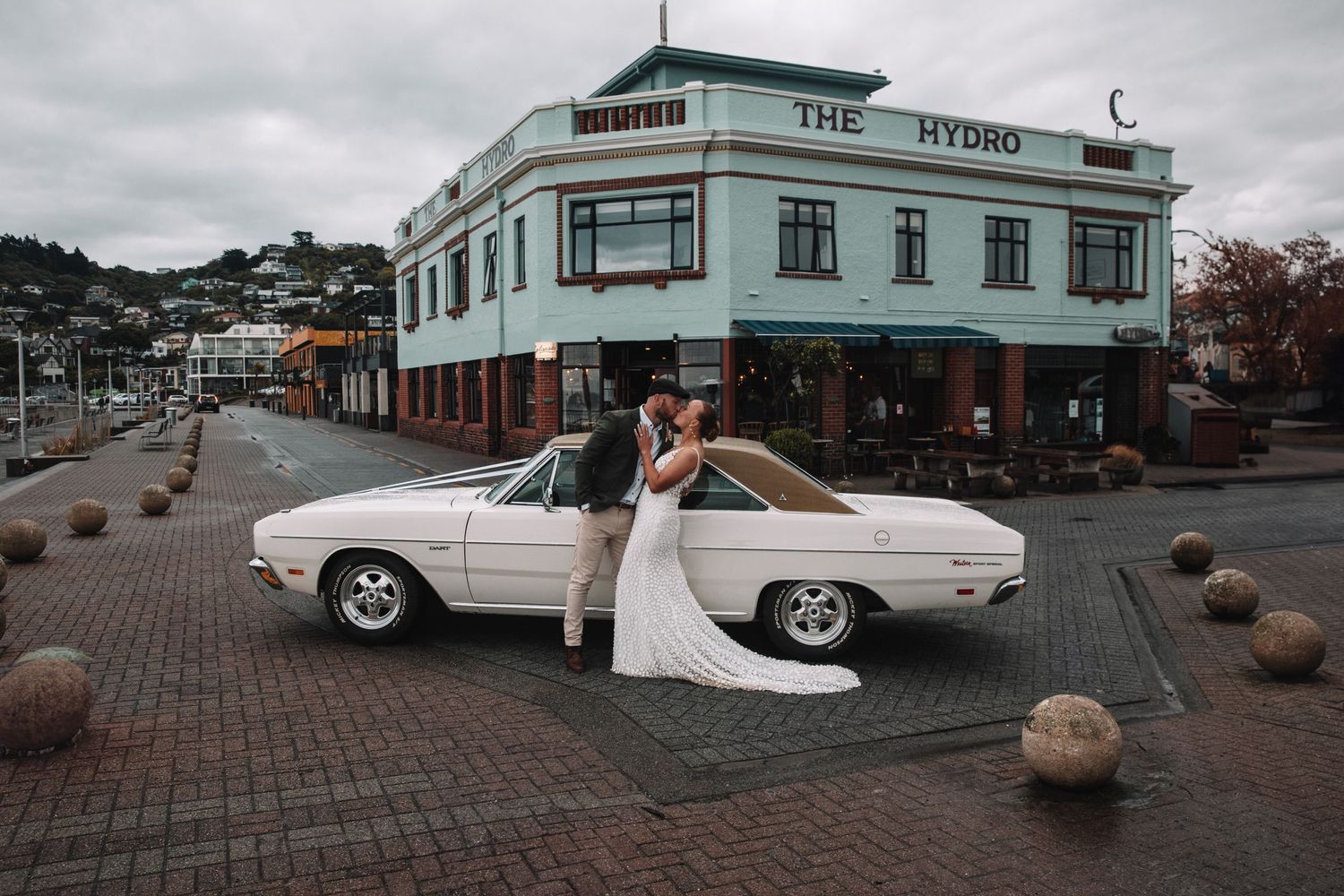 A wedding couple poses with a classic white car in front of a historic building.