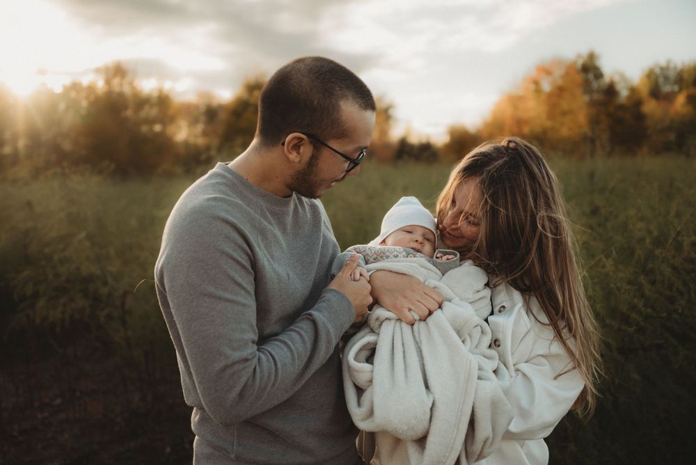 A couple lovingly holds their swaddled baby while standing in a golden-lit field at sunset.