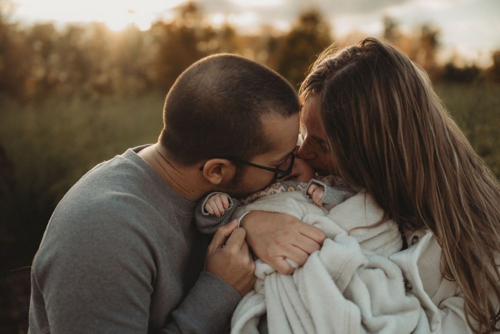 Parents share an intimate moment while embracing their newborn baby outdoors at dusk.
