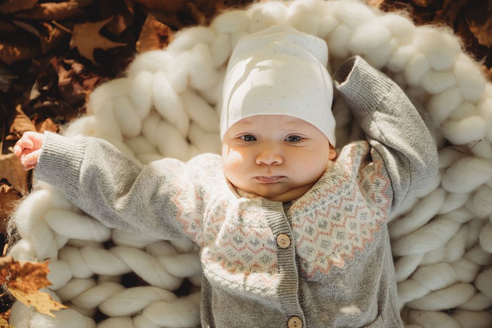 Baby wearing a white hat and gray sweater lies on chunky knit blanket surrounded by fall leaves.