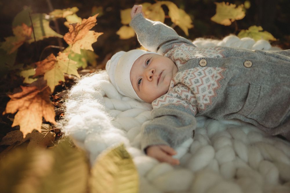 Infant in cozy winter attire rests on white knitted blanket with autumn leaves.