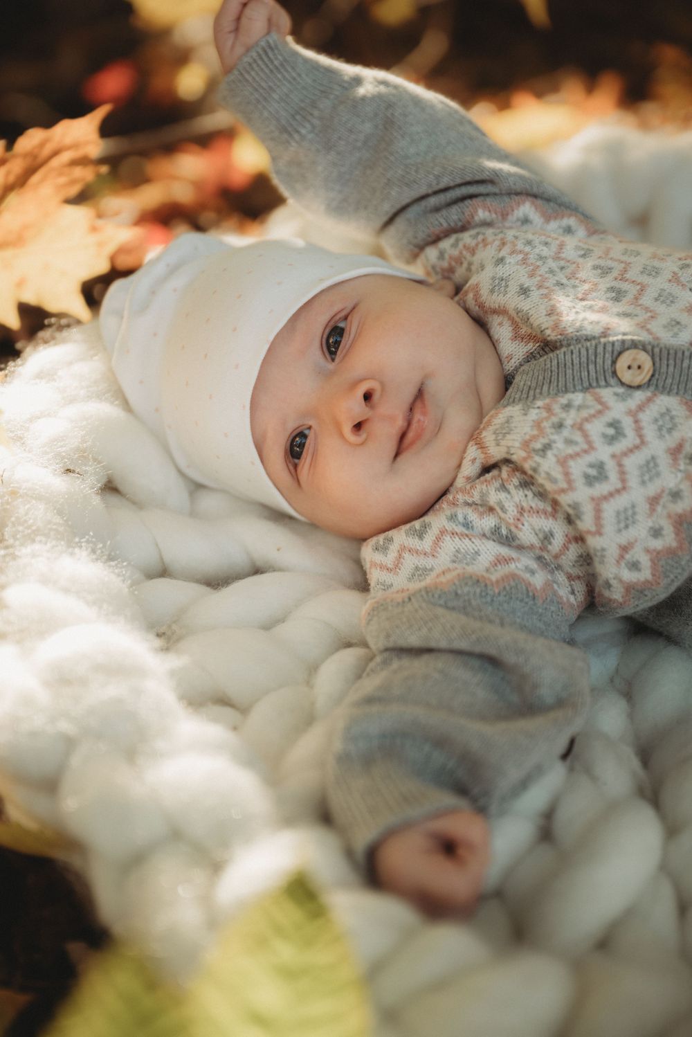 Baby in white hat and patterned sweater smiles while lying on chunky knit blanket.