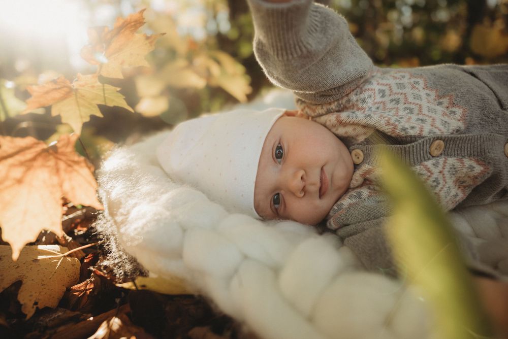 Baby in winter clothing rests on white blanket among fall foliage.