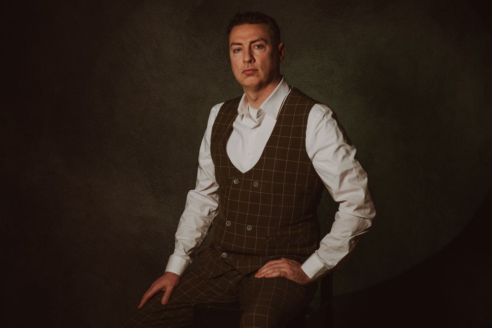 Professional studio portrait showing dark brown checkered vest and white dress shirt against moody backdrop.