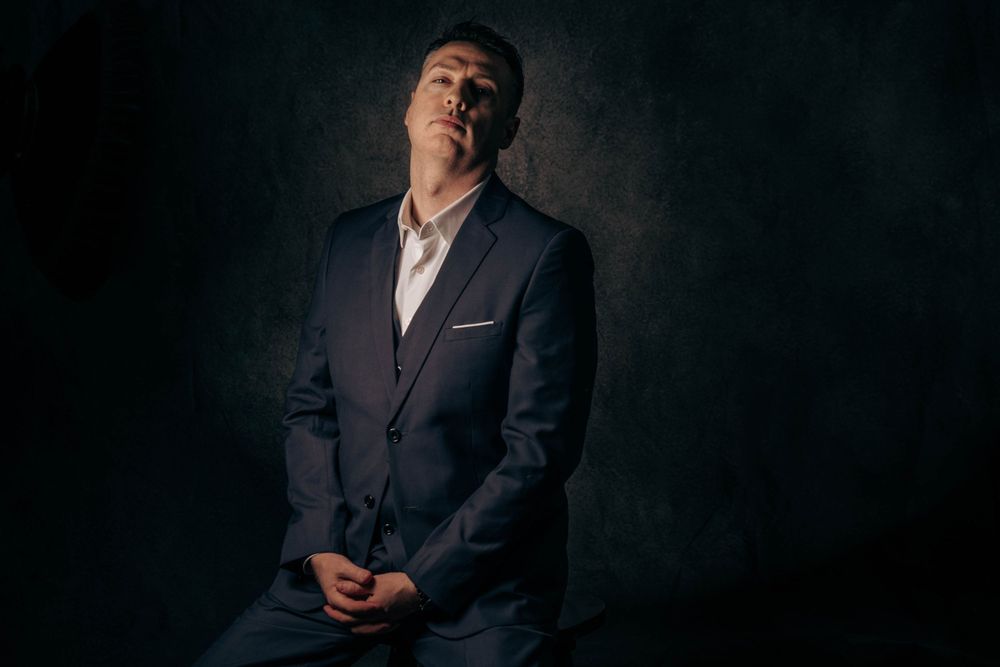 Dramatic low-key portrait featuring navy suit and white shirt against dark background.