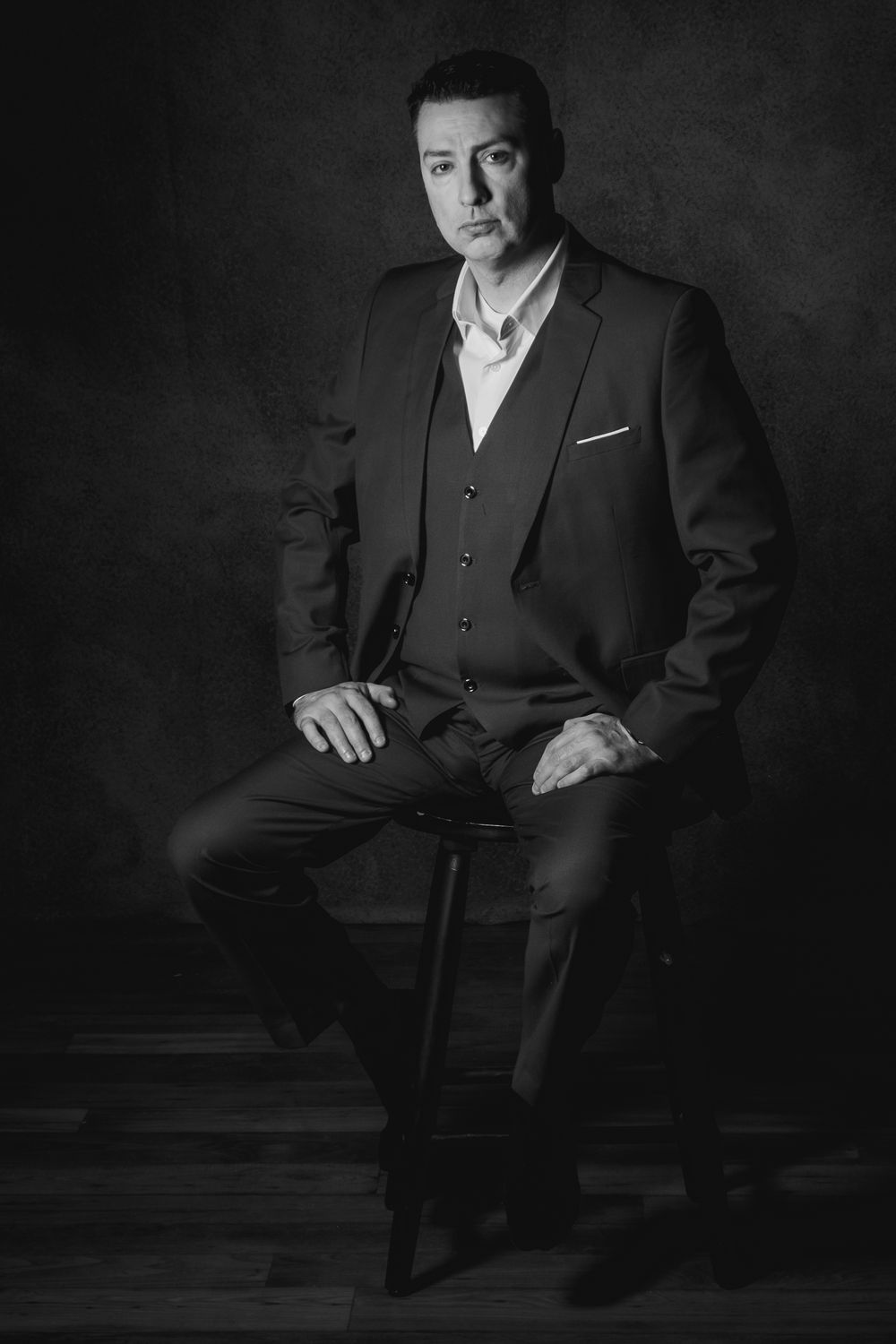 Classic black and white portrait of person in three-piece suit seated on stool.