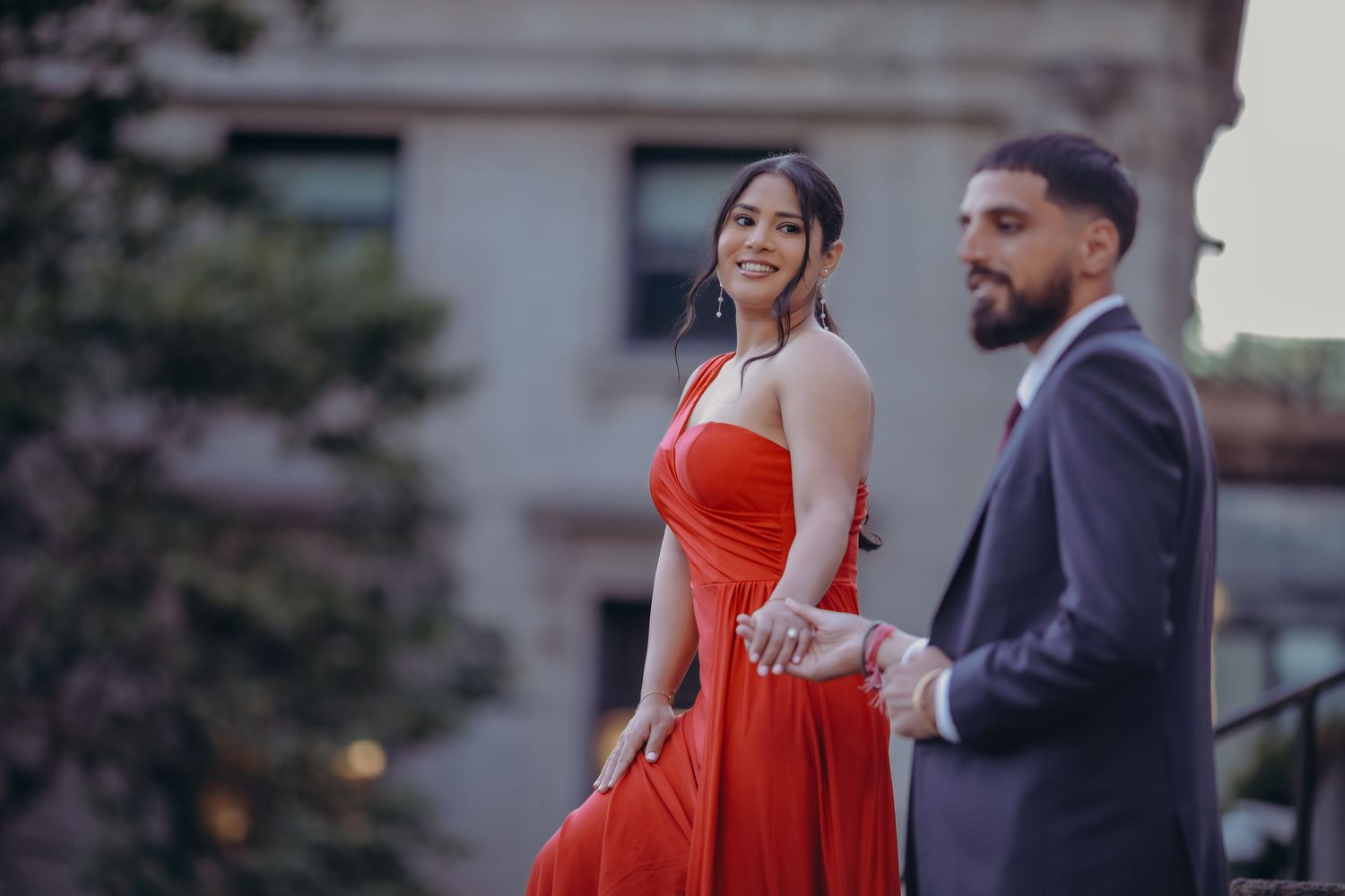 Couple photoshoot with bride wearing red dress and looking at the groom, with dark aesthetic background.
