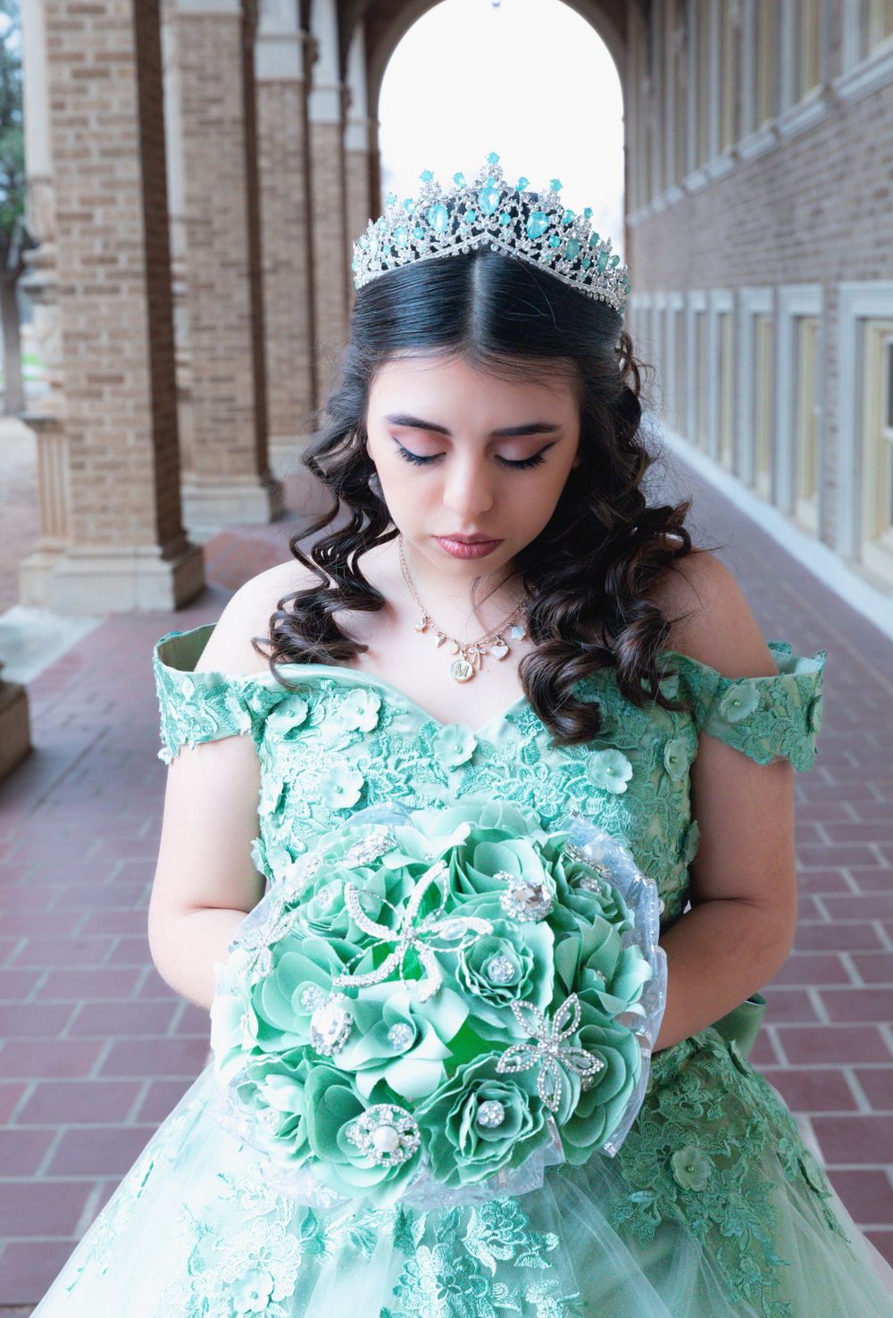 A person wearing a mint green off-shoulder ball gown with crystal tiara and holding a floral bouquet in an arched hallway.