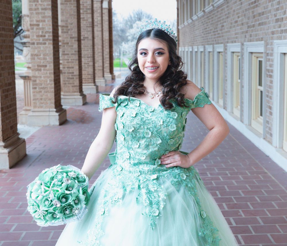 A person poses in an ornate mint green quinceanera dress with floral appliques and matching bouquet in a brick hallway.