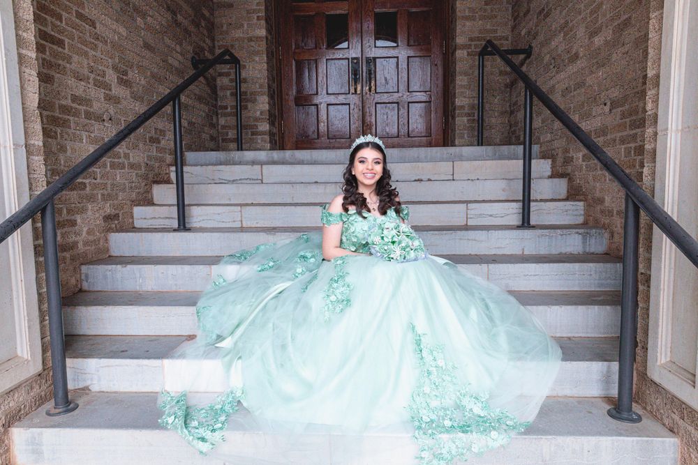 A mint green quinceañera dress with floral details worn by someone sitting elegantly on stone steps.
