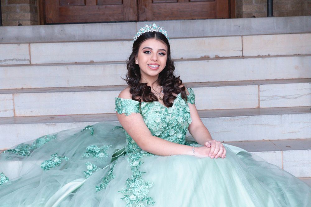 A person in a mint green quinceañera ball gown adorned with floral details poses elegantly on stone steps.