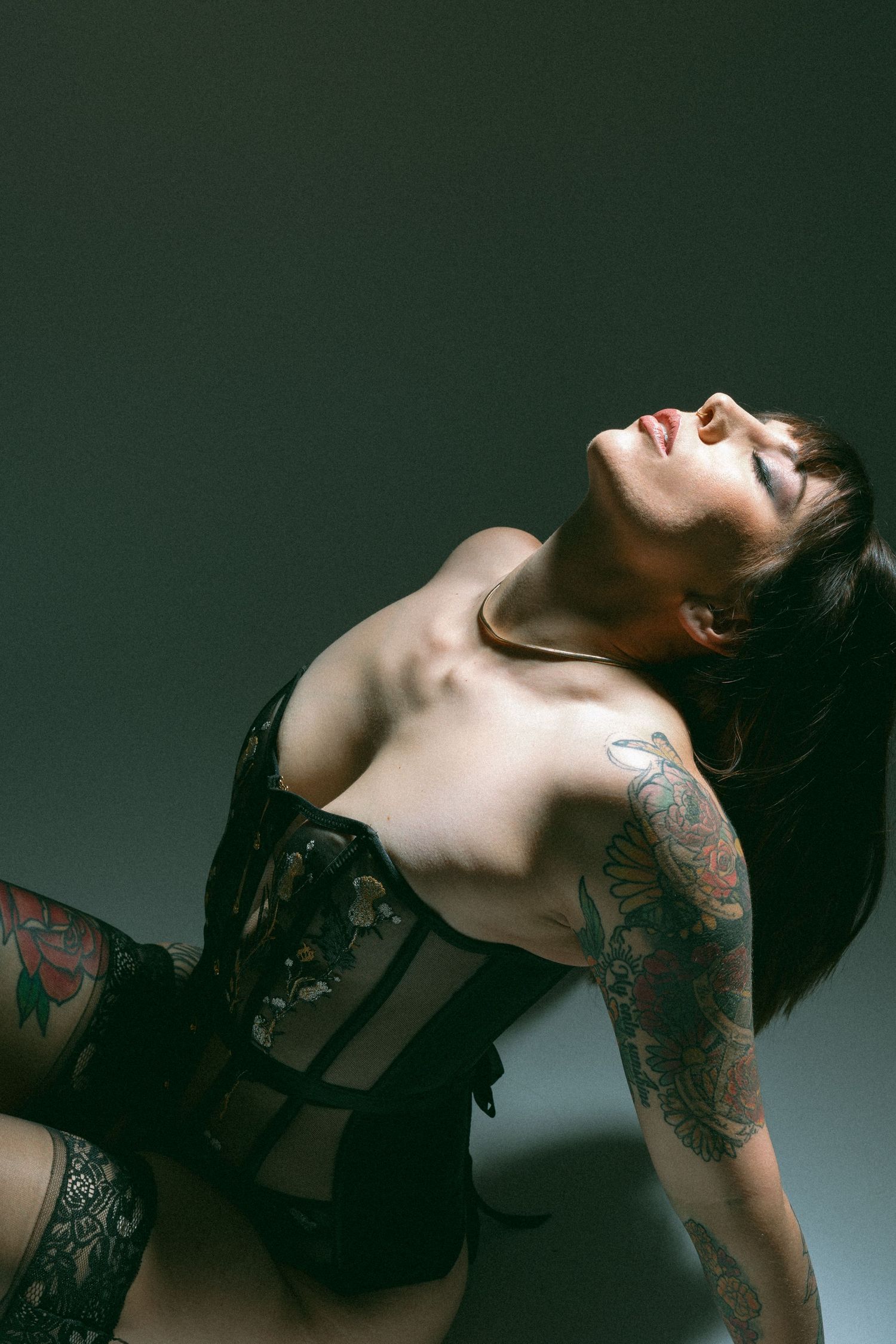 Woman in black lingerie arches back, displaying tattoos on arms in artistic pose against dark background.