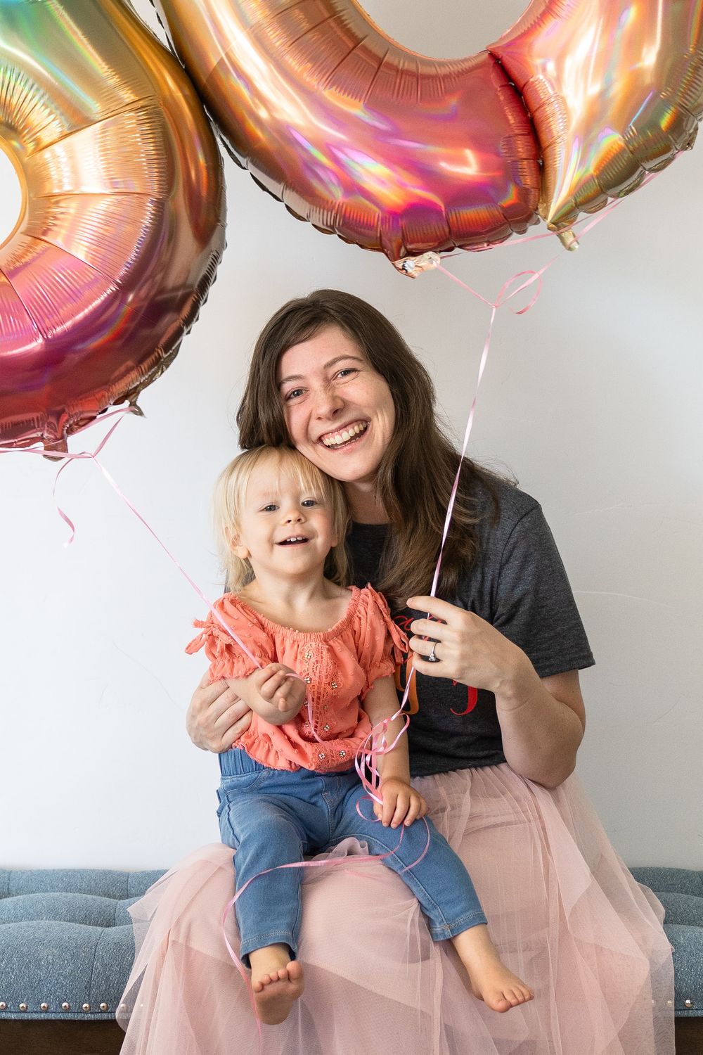 A mom and baby scene sit together on a blue couch wearing pink and coral holding balloons in Saratoga Springs, Utah.