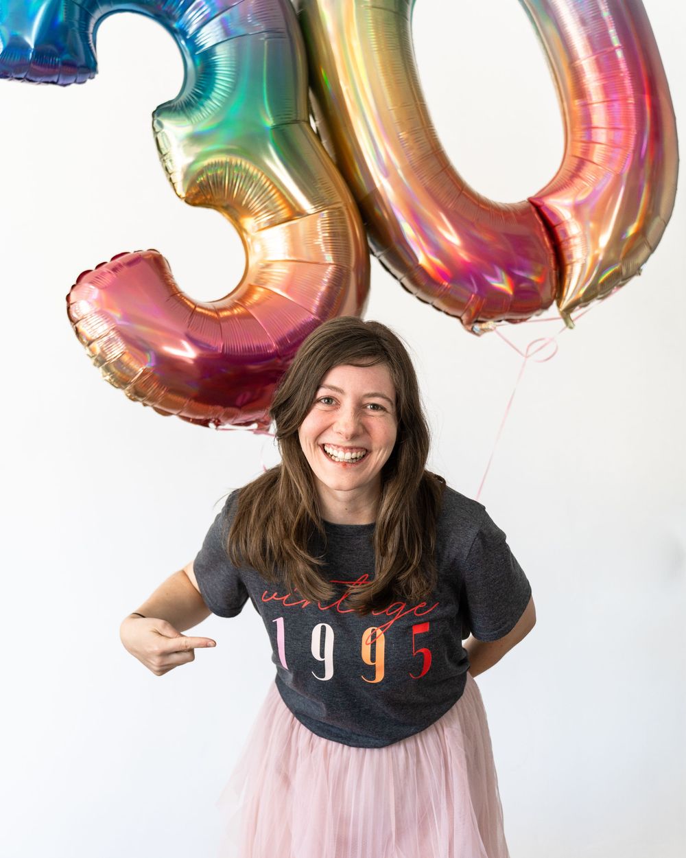 A photographer poses with rainbow-colored number 30 balloon against white background in Studio in Utah county, Utah.