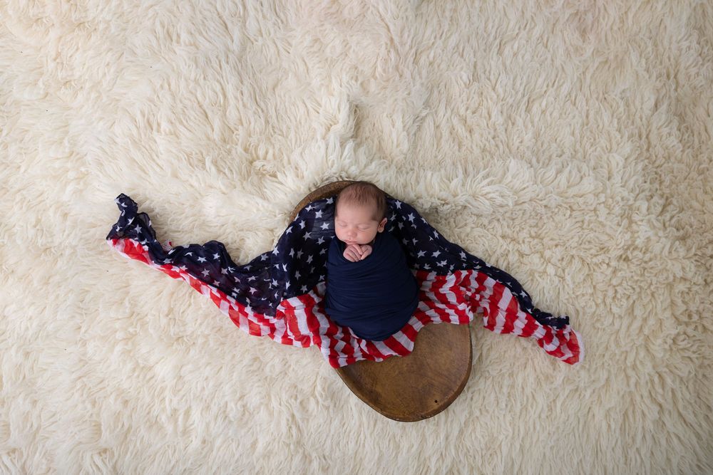 Sleeping newborn wrapped in American flag themed blanket laying on white carpet with penny.