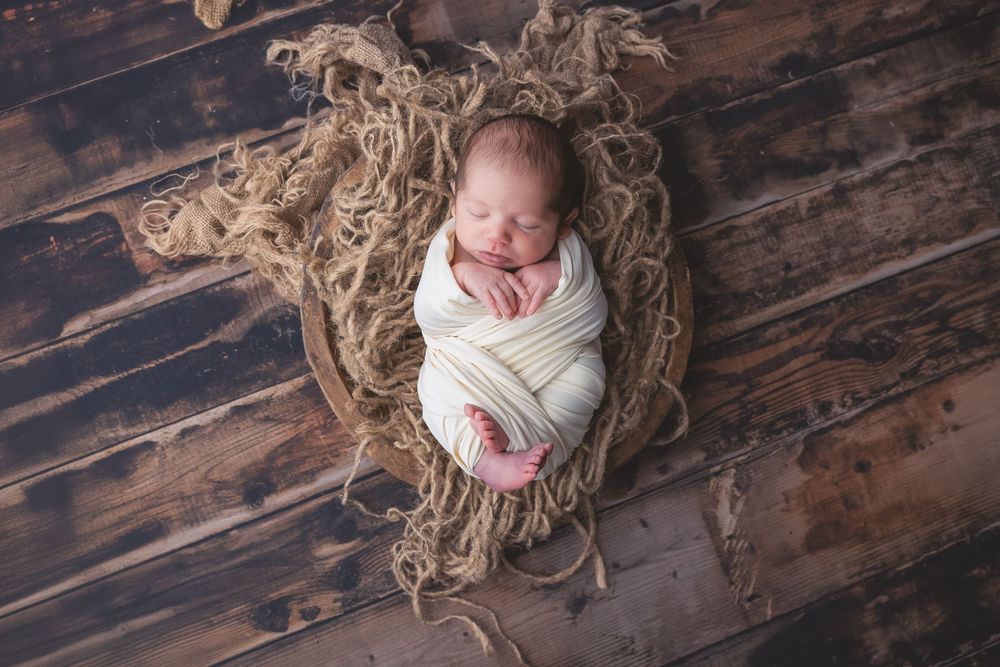 Sleeping newborn wrapped in cream swaddle blanket resting on straw bedding and rustic wood floor.