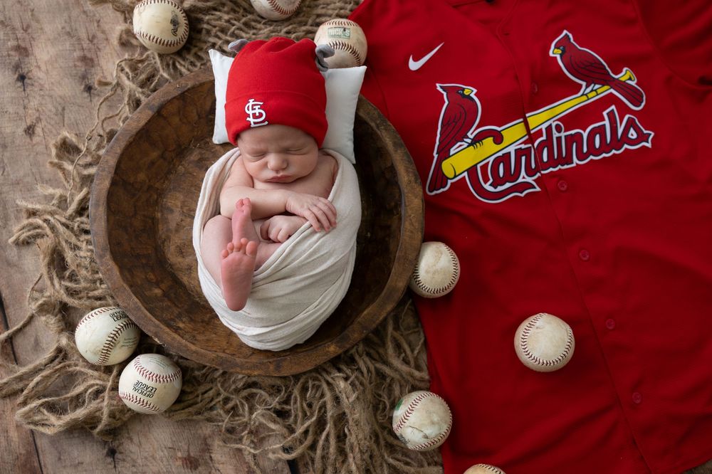 A newborn baby surrounded by baseballs and St. Louis Cardinals baseball gear in a wooden bowl.