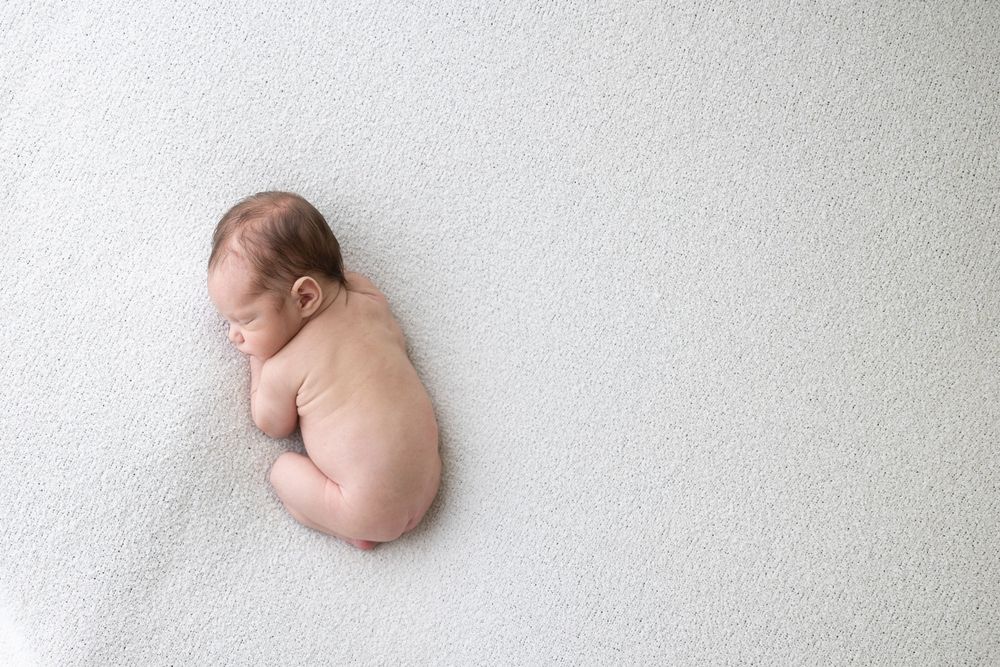 A sleeping newborn baby curled up on white blanket background during portrait session.