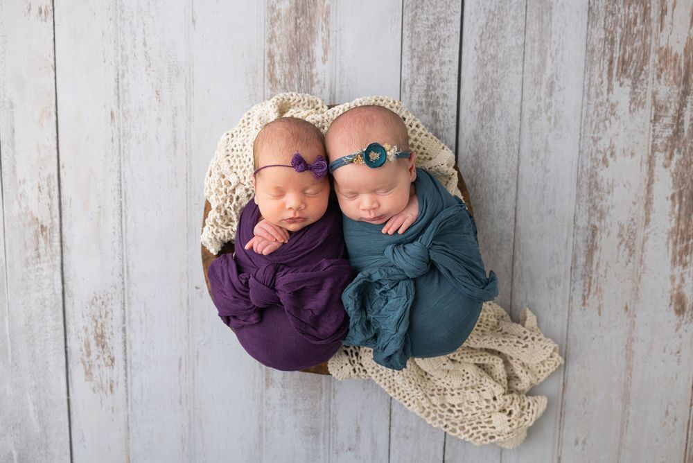 Two newborn babies wearing purple and blue wraps snuggle together on cream-colored blanket against rustic wood backdrop.