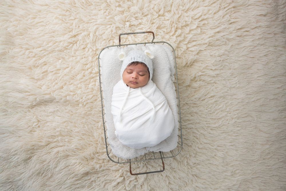 A swaddled newborn baby rests peacefully in a metal wire basket on a soft cream-colored carpet.