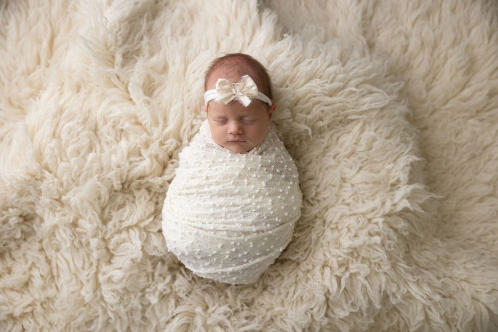 Newborn baby swaddled in white fabric lying on cream-colored fluffy fur backdrop wearing delicate headband.