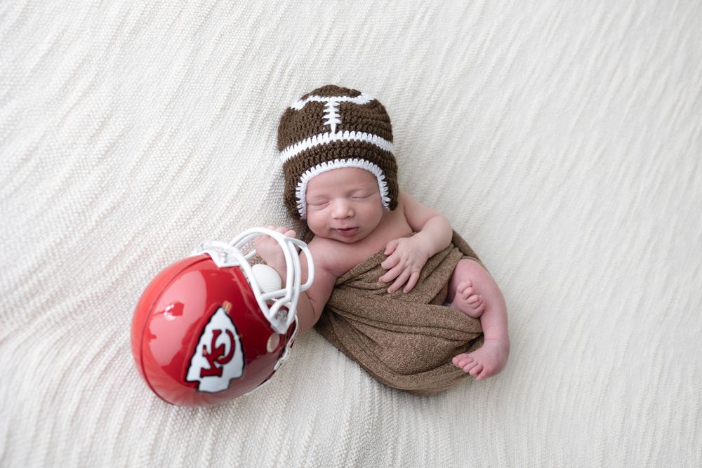 Newborn baby wearing football beanie lies sleeping next to Kansas City Chiefs football helmet on white carpet.