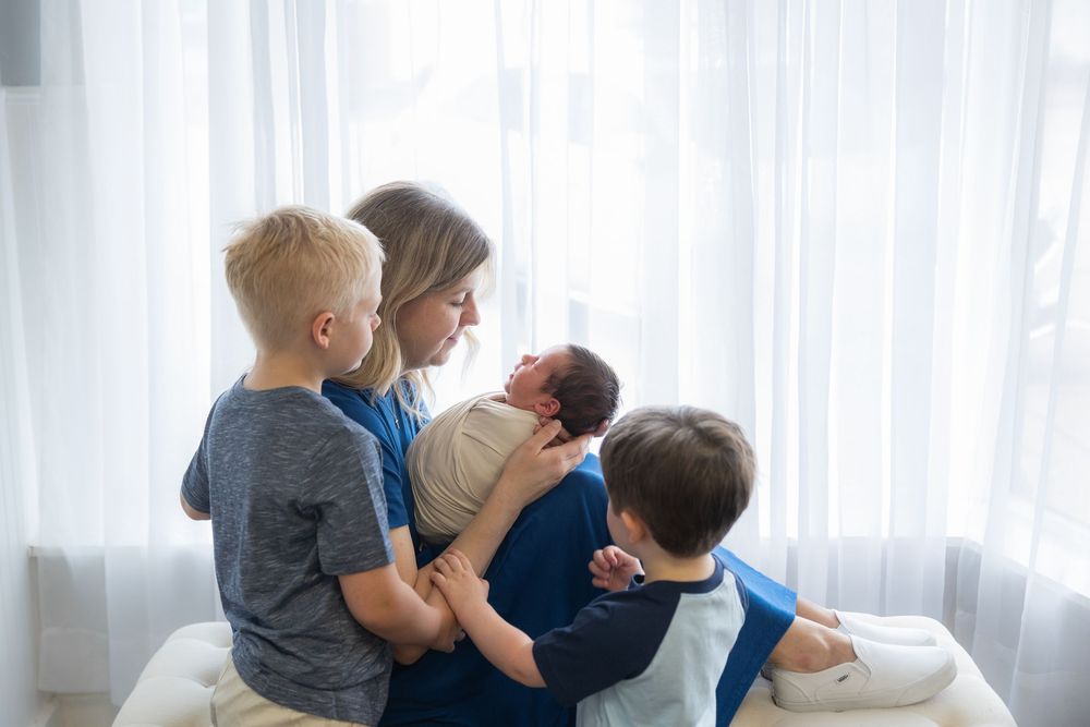 Family members sit together on white couch near window with sheer curtains while looking at newborn baby.
