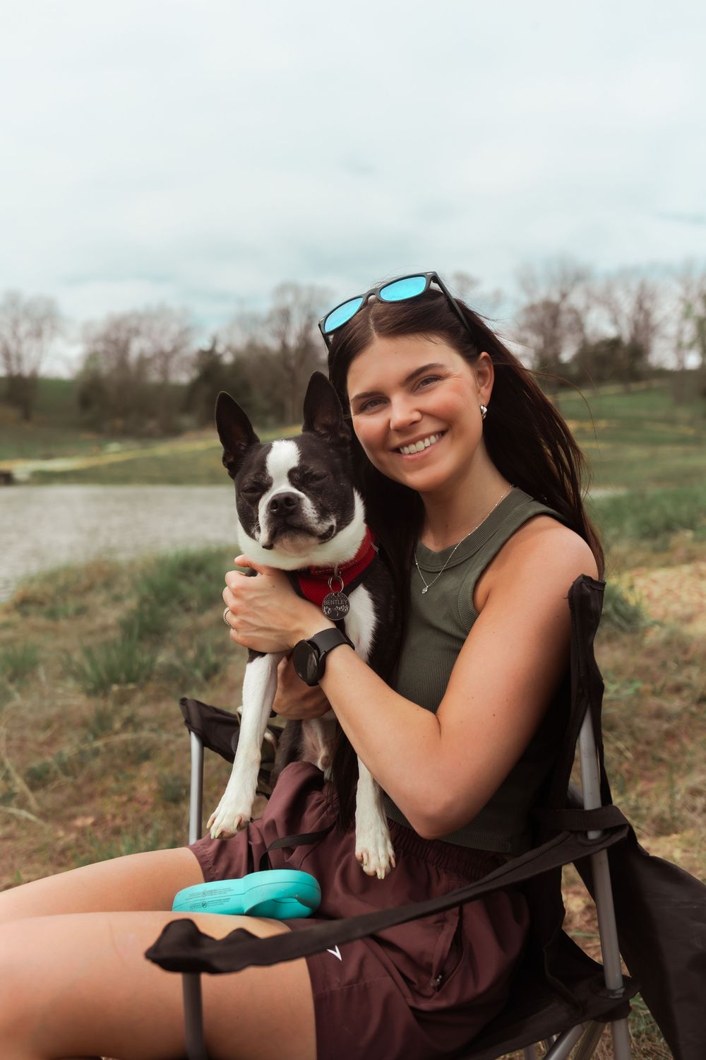 Boston Terrier puppy sits with companion on chair in outdoor field setting with sunglasses perched on head.