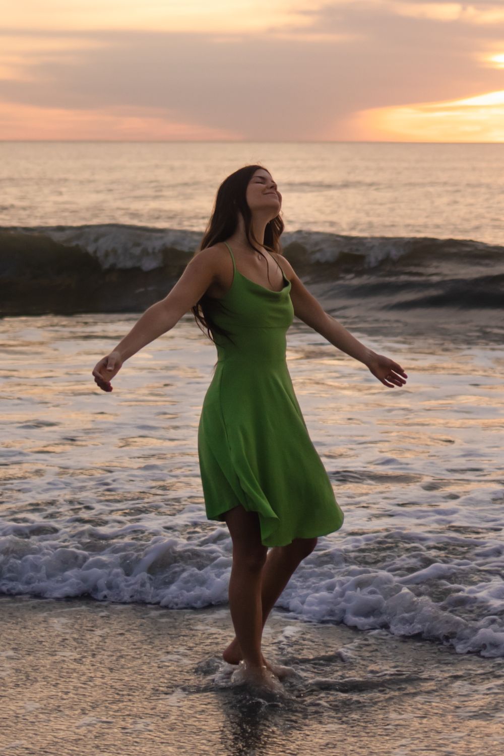 Person in flowing green dress twirls on beach at sunset as waves crash on shore in golden light.