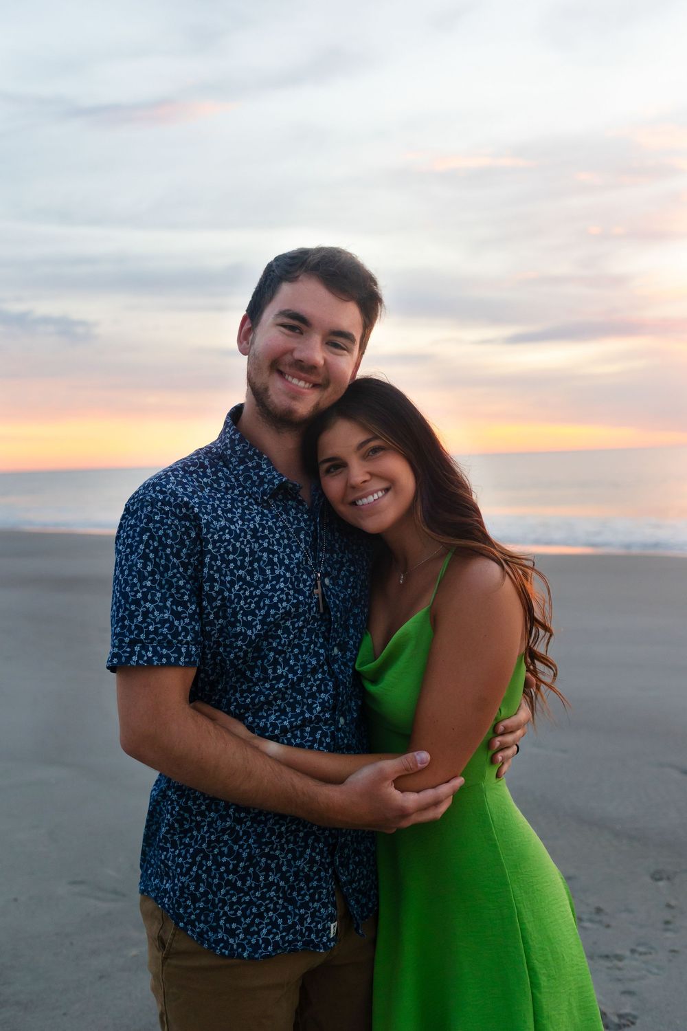 A couple embraces on a beach at sunset, with the person in green dress smiling warmly against the colorful sky.