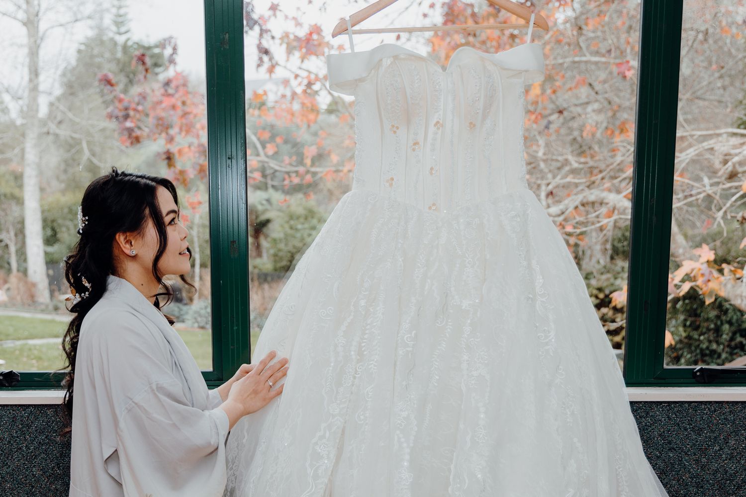 White wedding gown displayed in sunlit window with autumn foliage outside.