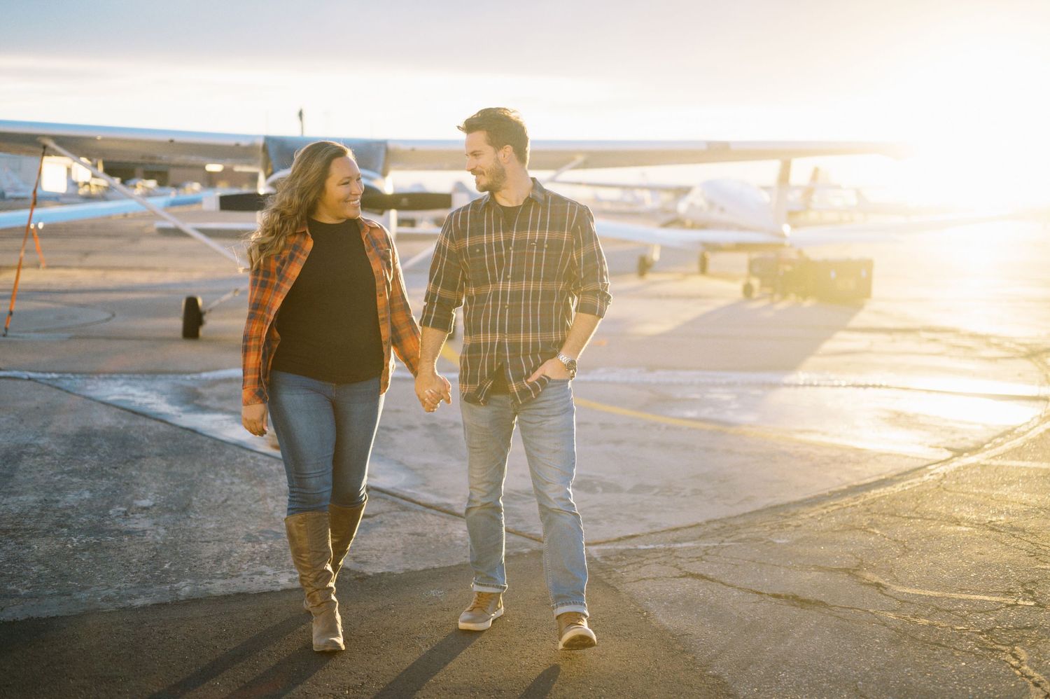 A couple walks hand in hand across an airport tarmac at sunset.