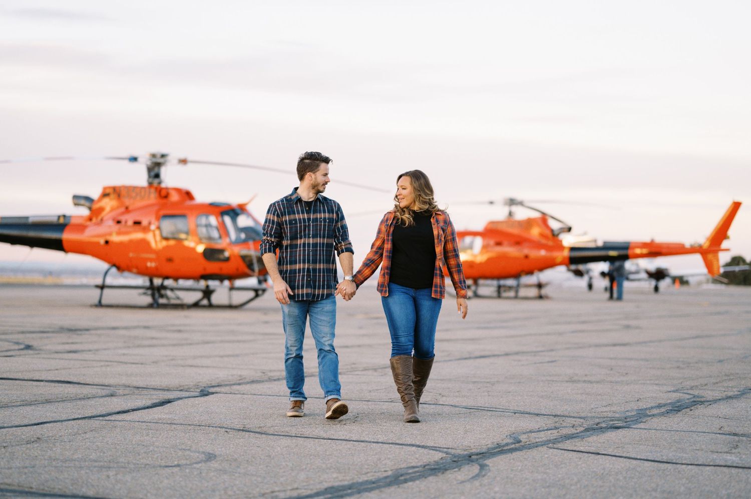 A couple walks together on an airport tarmac with orange helicopters in the background.